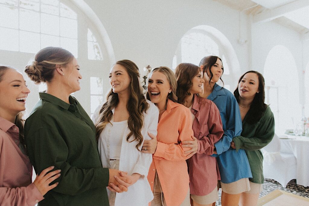 Bride smiles towards bridesmaids who are dressed in different colored oversized button up shirts.