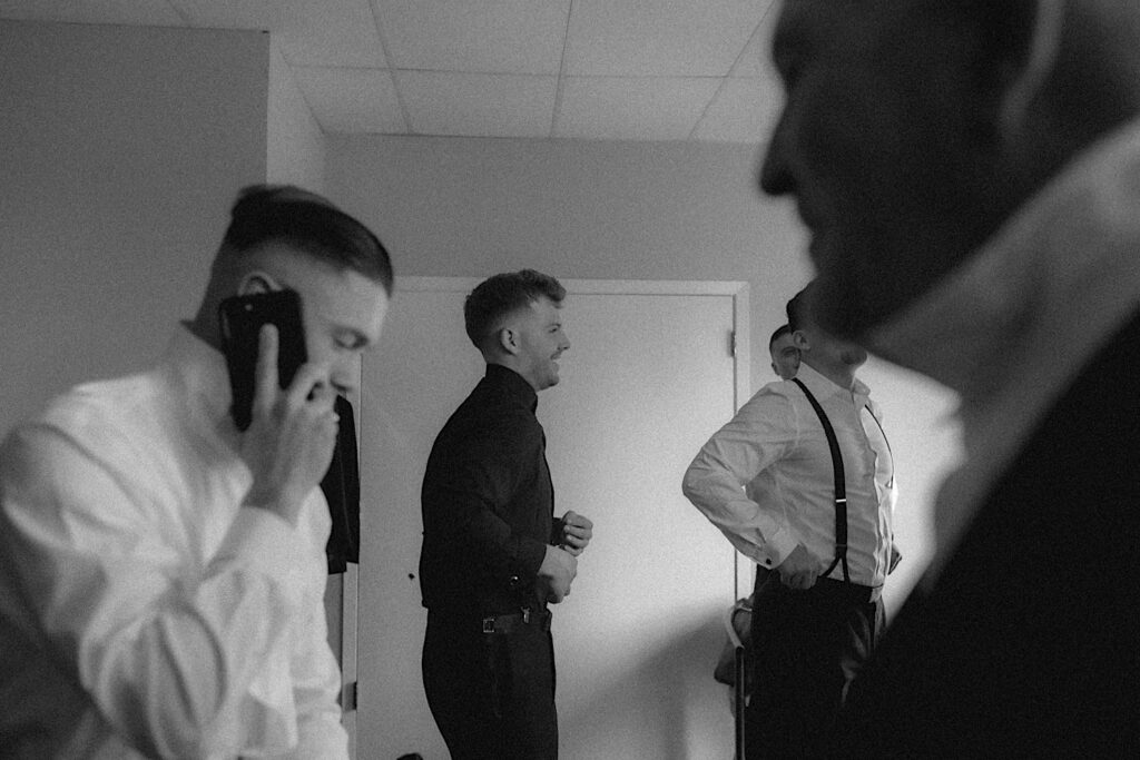 Groomsmen all get ready in room in filmy black and white photograph before wedding ceremony. 