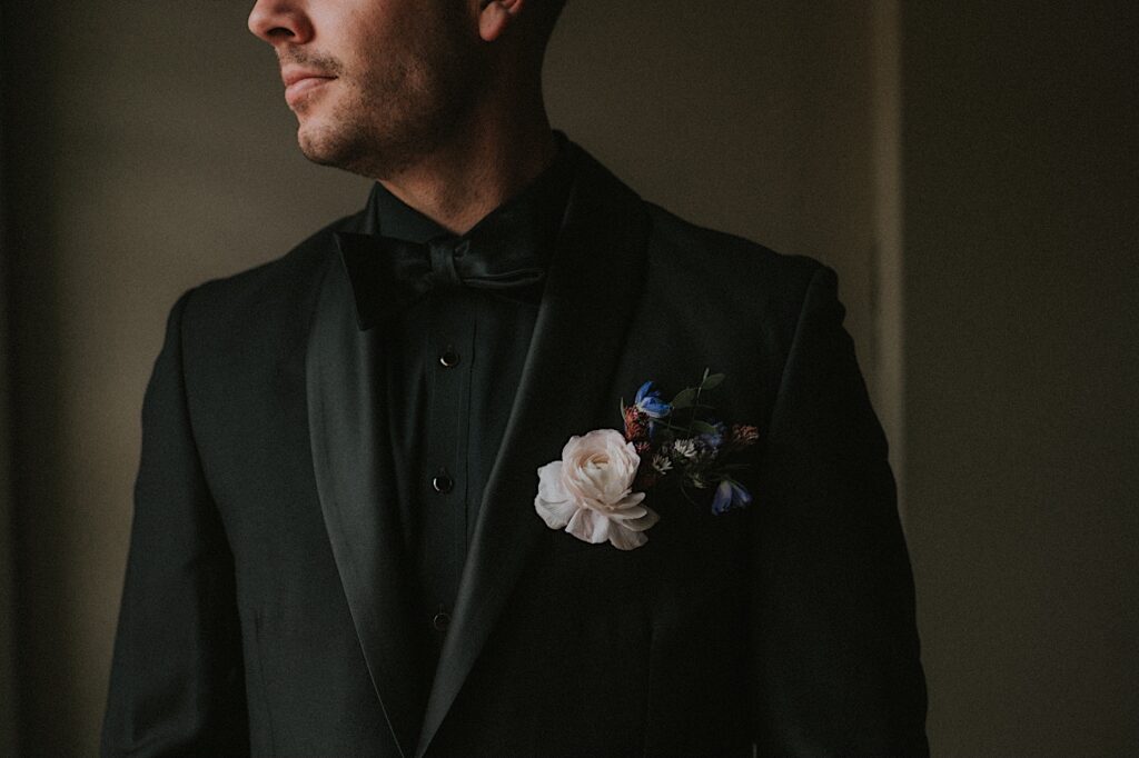 Groom stands in all black suit and black bow tie with rose boutonnière.  