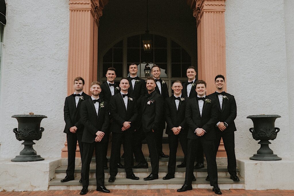 Groom stands with groomsmen in all black tuxes on steps of Ravisloe Country Club between two cement urns. 