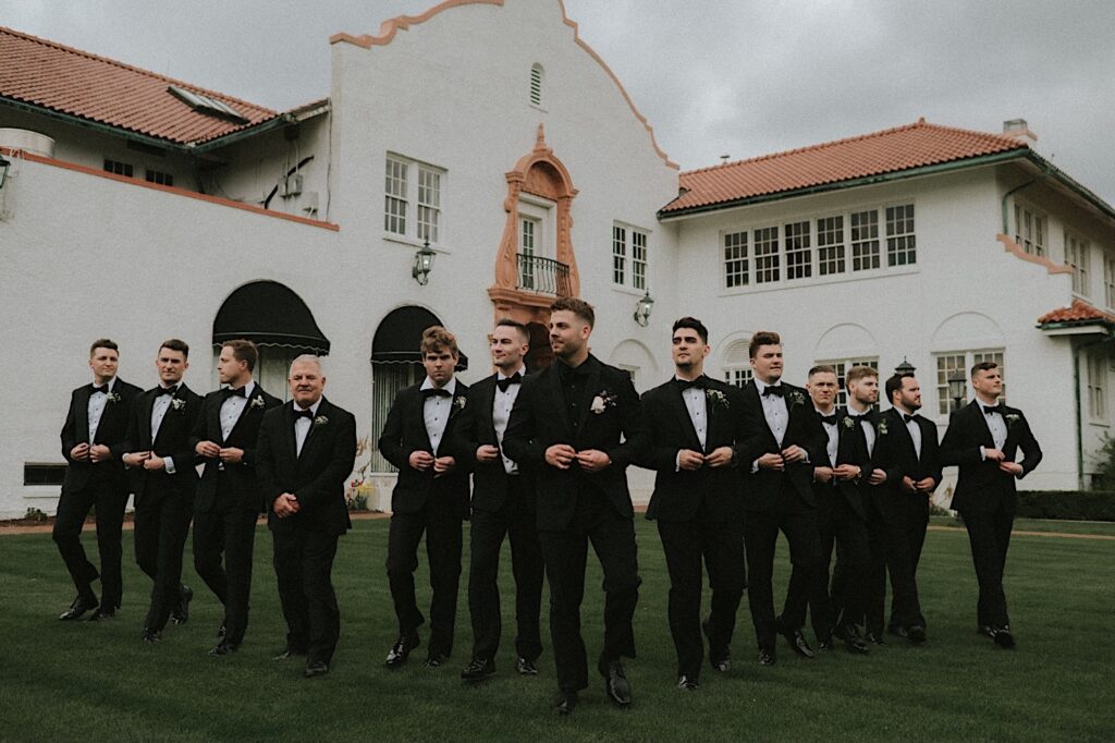 Groomsmen walk behind groom in a v-shape with Ravisloe Country Club behind them in the background. 