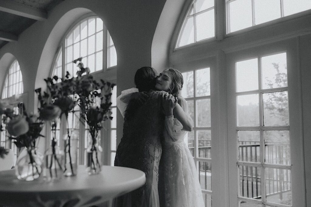 Bride hugs her mom in front of beautiful windows before wedding ceremony. 