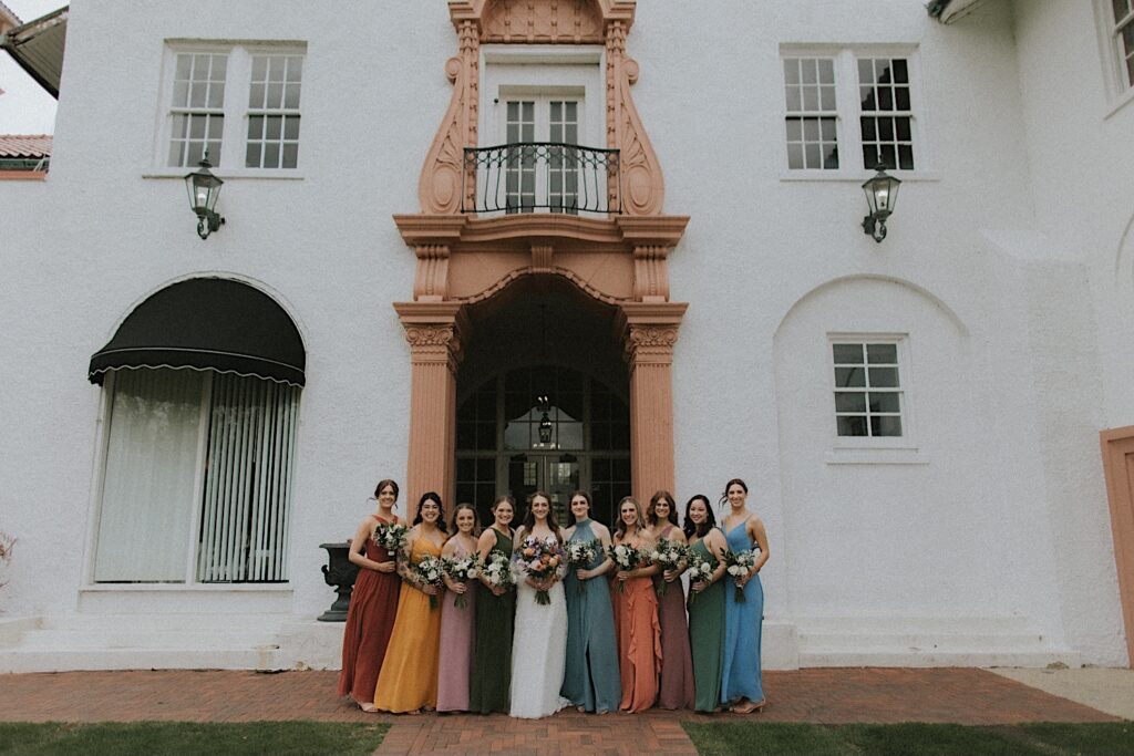 Bride poses with her bridesmaids in different colored dresses in front of entrance at Ravisloe Country Club. 