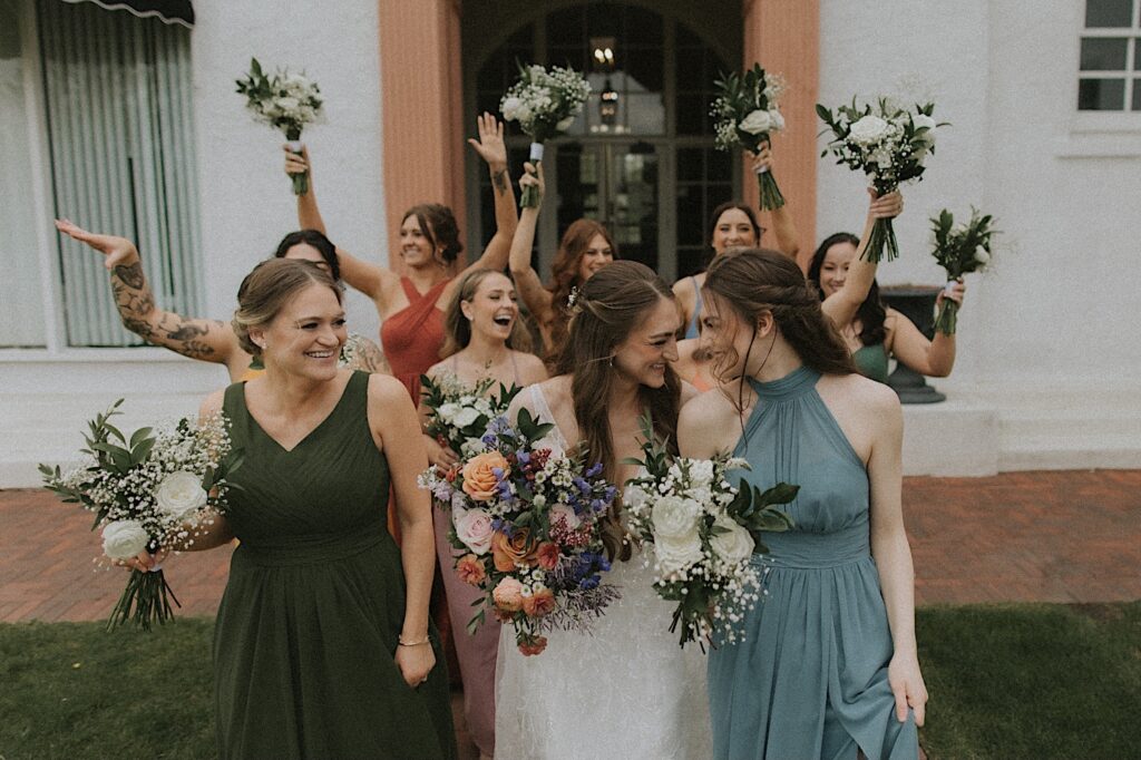 Bride leans into bridesmaid smiling while there are bridesmaids standing behind them holding their white bouquets in the air. 