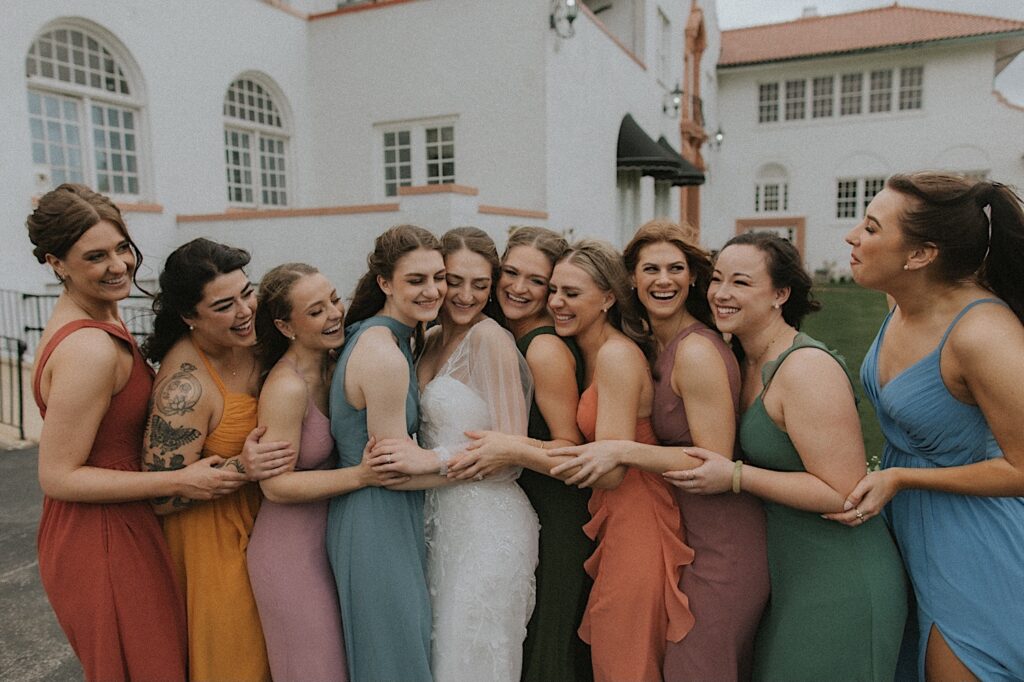 Bridesmaids in different colored dresses all lean in to hug bride smiling and laughing with Ravisloe Country Club in the background. 