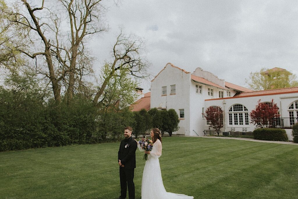 Bride stands behind groom before surprising him with a first look with Ravisloe Country Club in the background. 