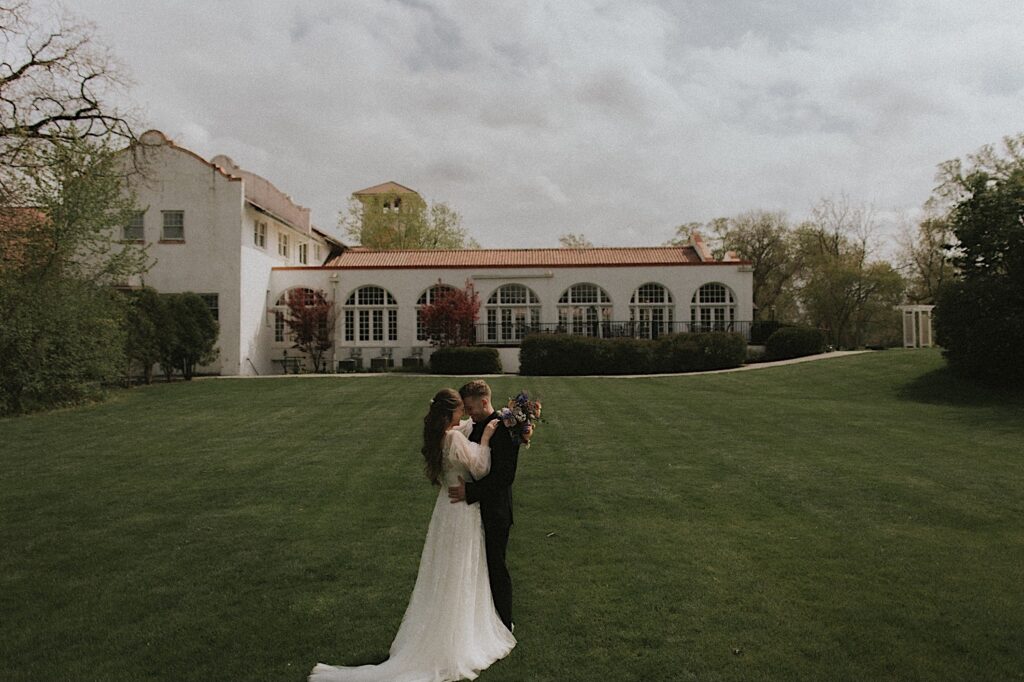 Bride and groom embrace and hold each other in grassy yard with Ravisloe Country Club in the background. 