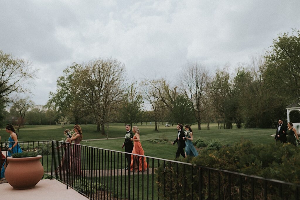 Groomsmen and bridesmaids make their way to the ceremony walking down a brick path with trees and grass in the background. 