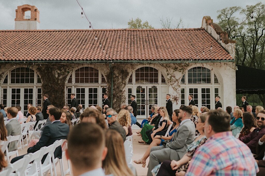 Groomsmen walk while the guests are seated in front of them. In the background is the White Ravisloe Country Club building covered in vines. 