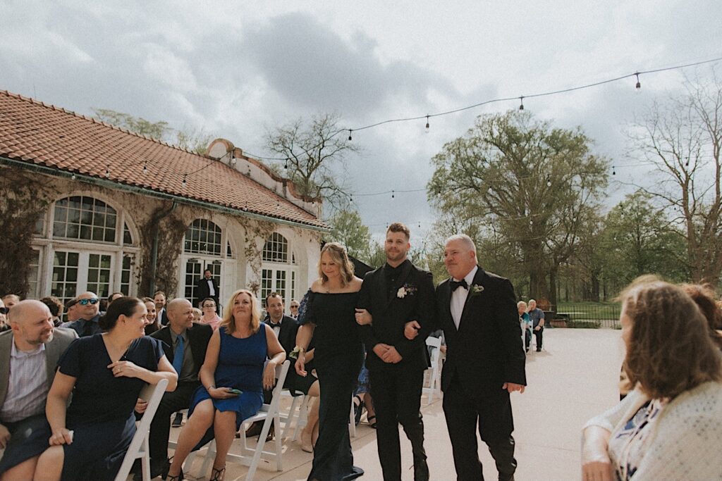 Guests watch as the Groom walks down the aisle with his parents. String lights are above them and the country club is on their left side. 