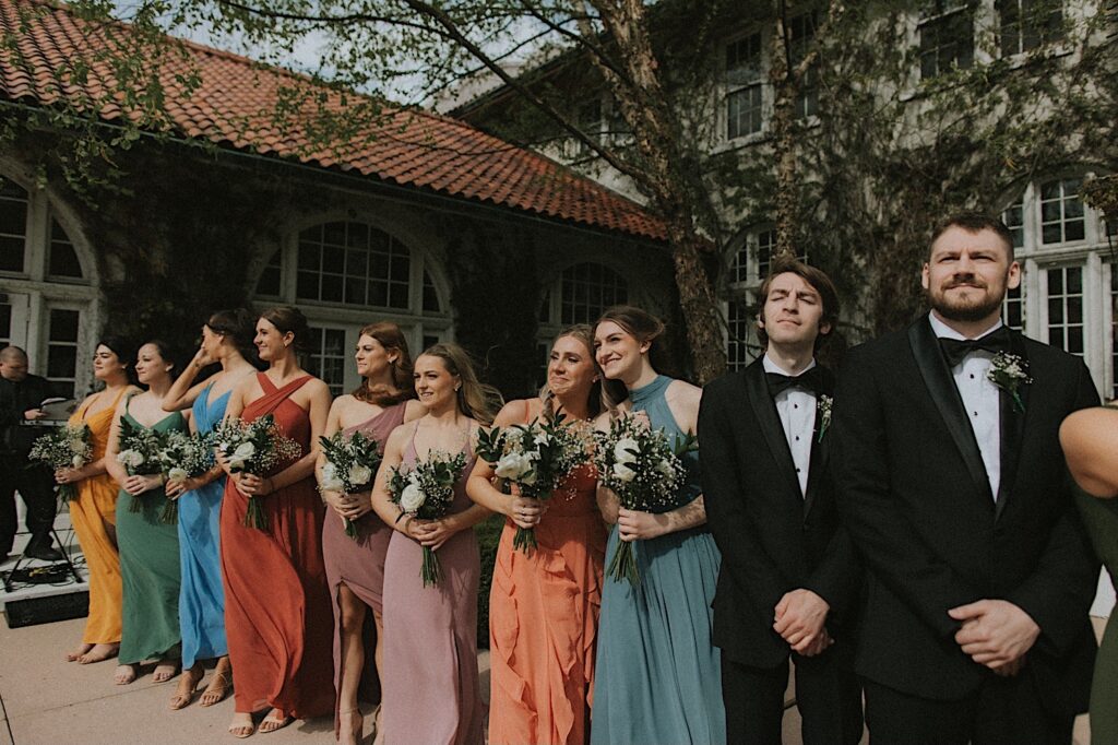 Bridesmaids in different colored dresses lean into each other and smile emotionally as the bride walks down the aisle. Ravisloe Country Club covered in vines stands behind them. 