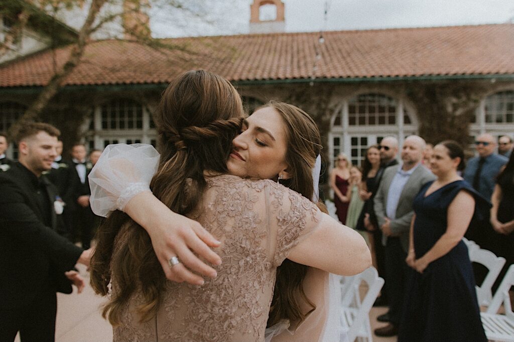 Bride hugs her mom at the end of the aisle before wedding ceremony starts as guests stand around them. 