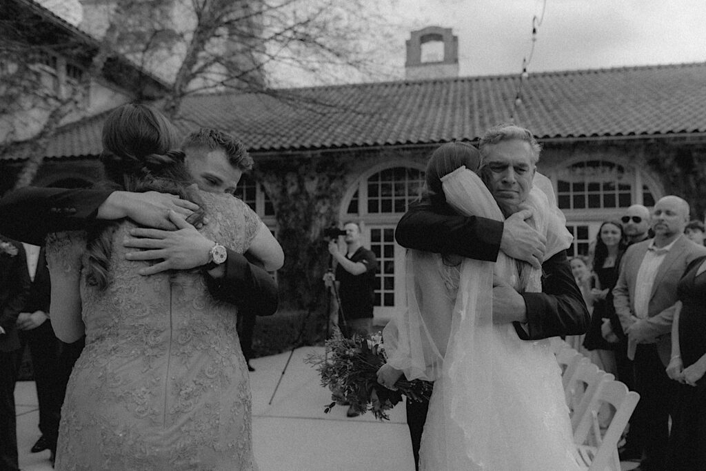 Bride hugs her father and the groom hugs Bride's mother at the end of the aisle as guests stand watching during the ceremony with Ravisloe Country Club in the backgrounnd. 