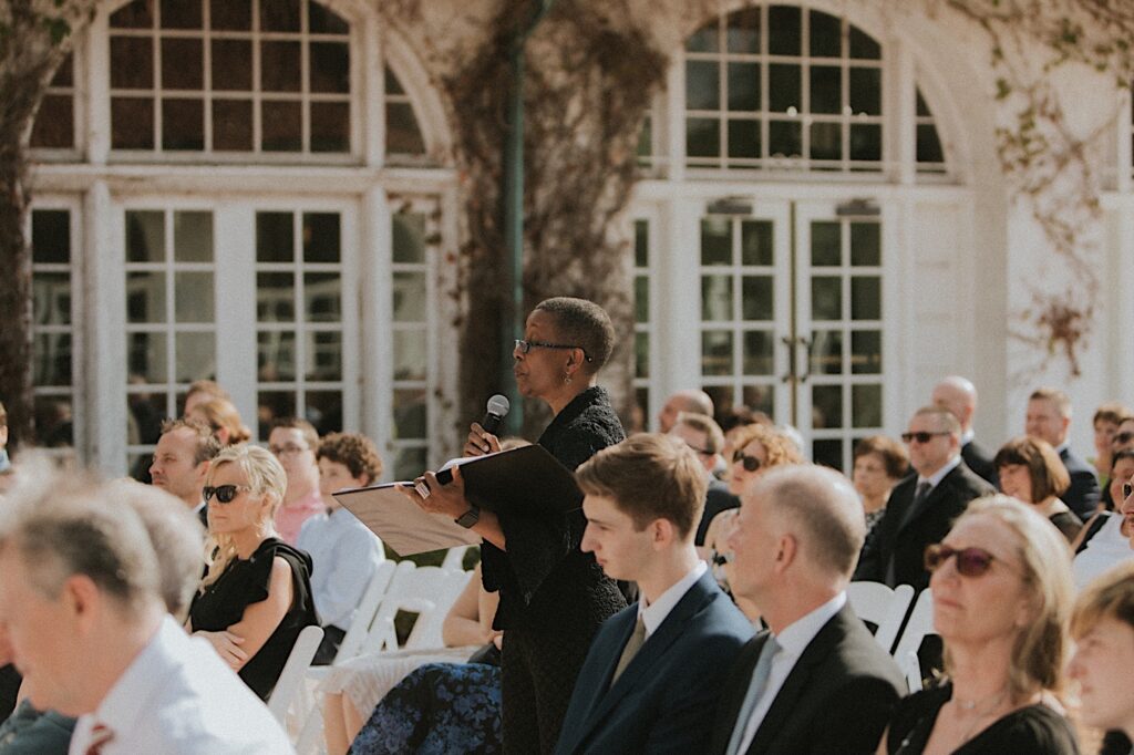 Wedding officiant makes her way down the aisle with a microphone in hand as guests look towards the front of the aisle. 