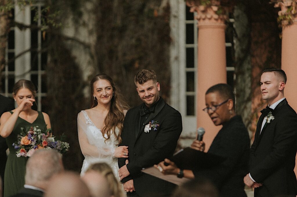 Bride and Groom stand arm and arm as the officiant speaks during wedding ceremony. In the background you can see the windows and peach columns of the Ravisloe Country Club. 