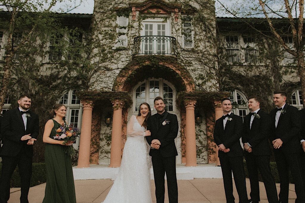 Bride and Groom stand arm in arm in front of the gorgeous entrance of Ravisloe Country Club smiling during wedding ceremony. 