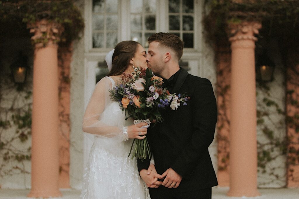 Bride holds colorful bouquet up to her face as her and groom kiss during wedding ceremony in front of entrance of Ravisloe Country Club. 