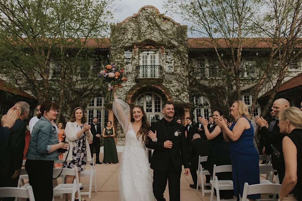 Bride holds up her bouquet in celebration as she and the groom make their way down the aisle after wedding ceremony. Their guests smile and cheer around them. 