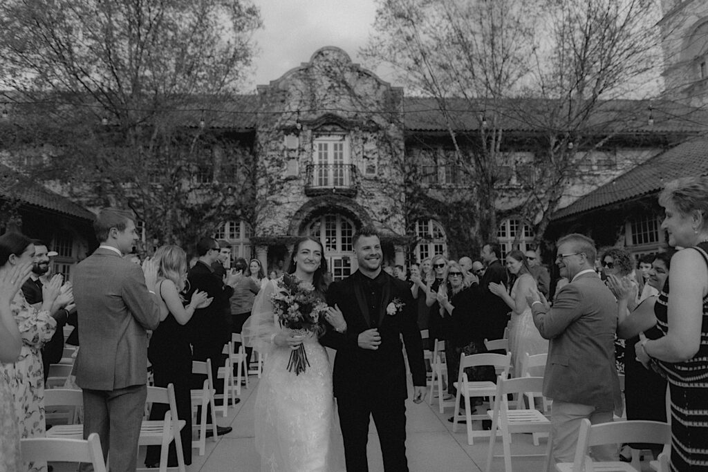 Black and White Photo of bride and groom walking down the aisle after wedding ceremony with guests cheering and clapping around them. 