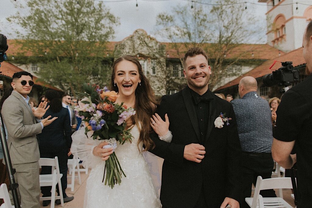 Bride holding a colorful bouquet and Groom in an all black tux smile as they walk arm in arm down to the end of the aisle. 