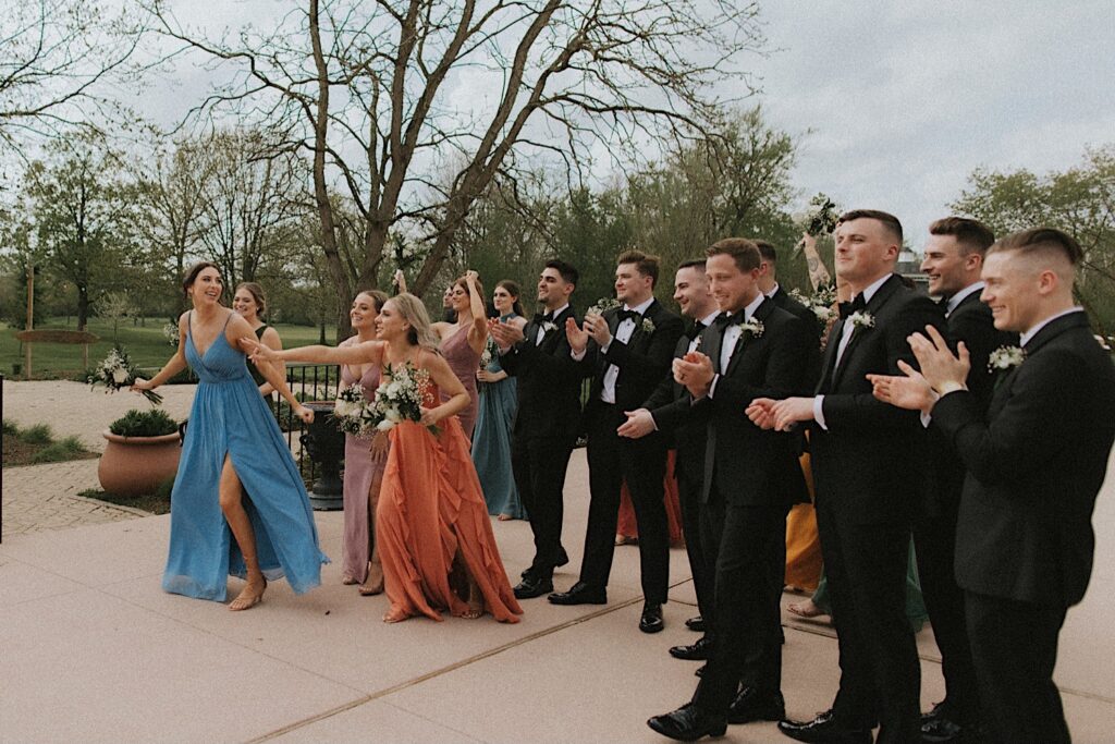 Bridesmaids and groomsmen clap as they run towards the bride and groom standing on pavement at the end of the aisle. 