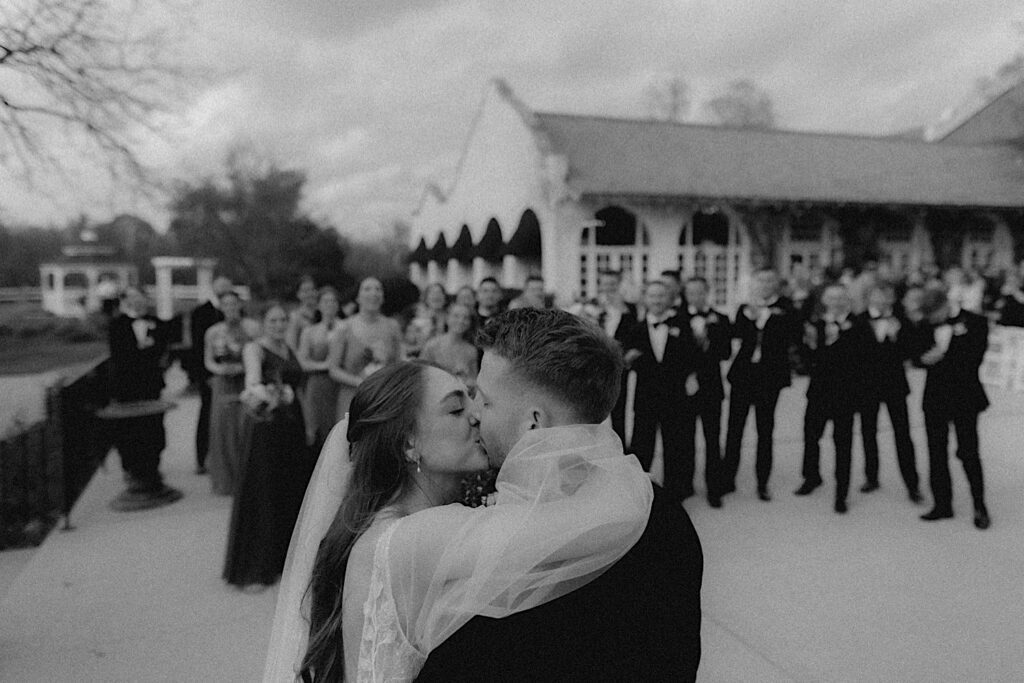 Black and White photograph of bride and groom kissing as their bridesmaids and groomsmen clap behind them. 