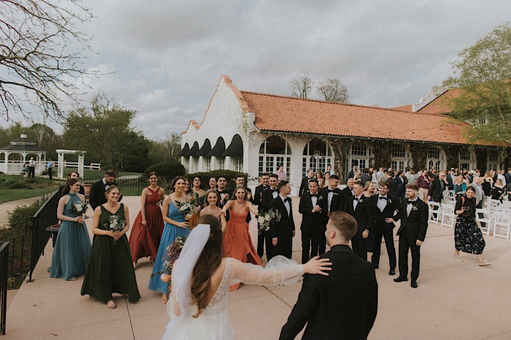 Bride and Groom run towards their wedding party as guests file out of their seats after wedding ceremony. In the background is the gorgeous Ravisloe Country Club and gazebos. 