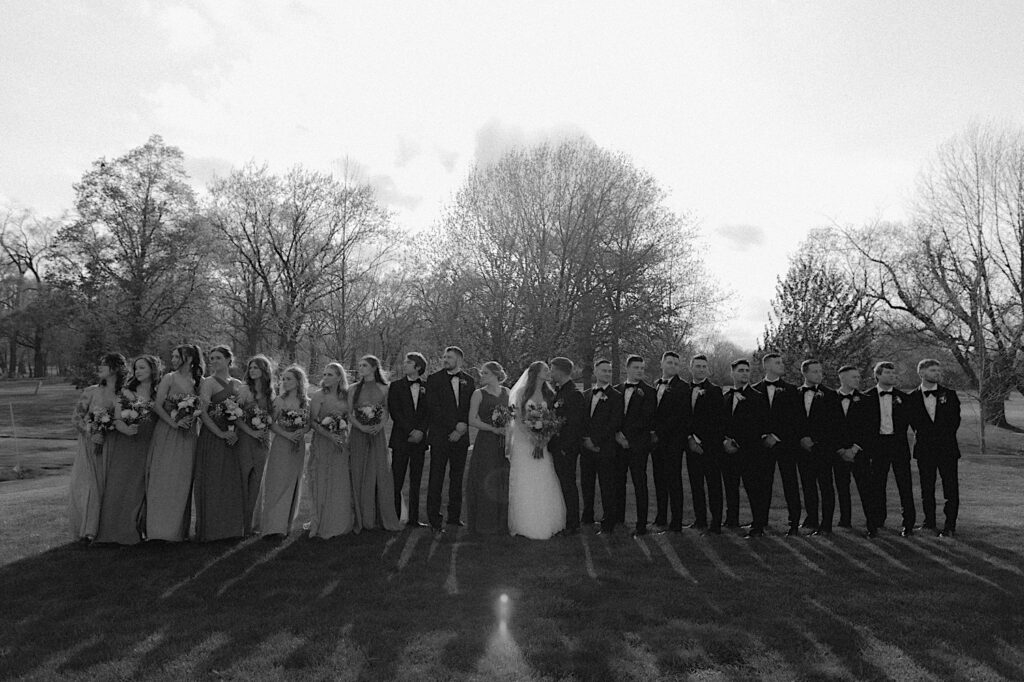 Wedding party stands out on the grass. The bridesmaids look left and the groomsmen look right as the bride and groom kiss in the middle. 