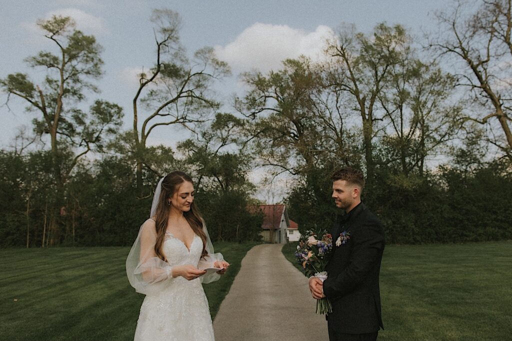 Bride and Groom stand on a sidewalk with the Ravisloe Country Club beyond a tree line as they read their intimate vows to each other. 