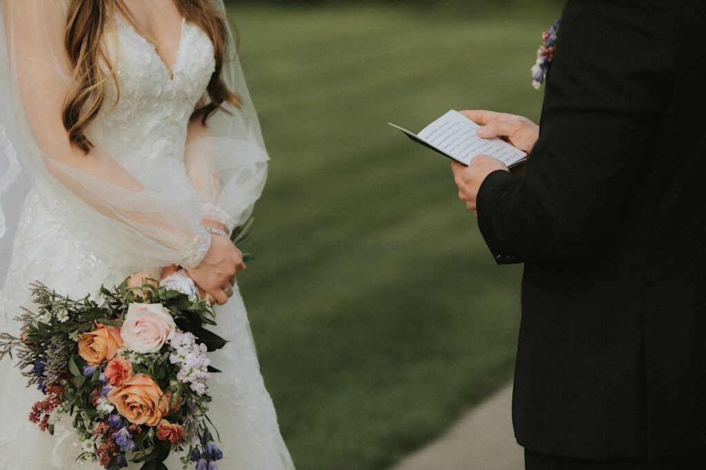 Chicagoland Wedding Photographer captures groom's hands holding book he wrote his vows in and bride's hands clasped holding her bouquet as she listens. 