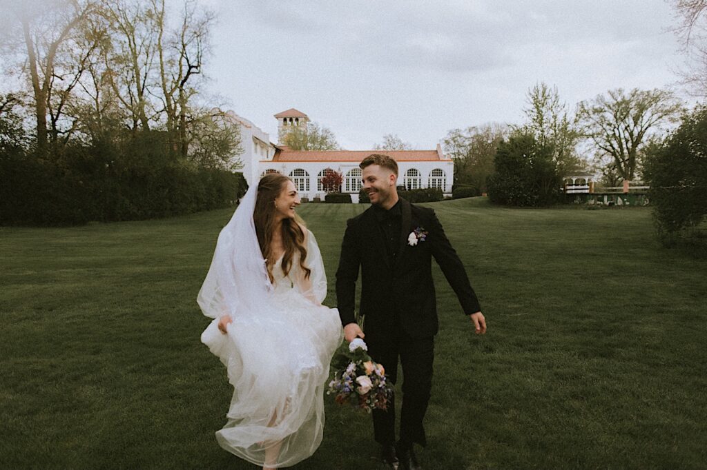 Bride and Groom hold hands as they run in the grass towards the camera smiling at each other with the Ravisloe Country Club in the background. 