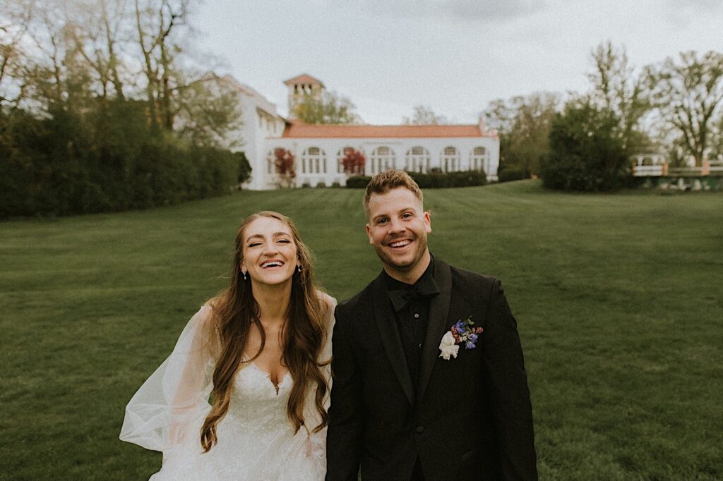 Bride and Groom smile smile big at the camera as they stand in the grassy lawn with the Ravisloe Country Club in the background. 