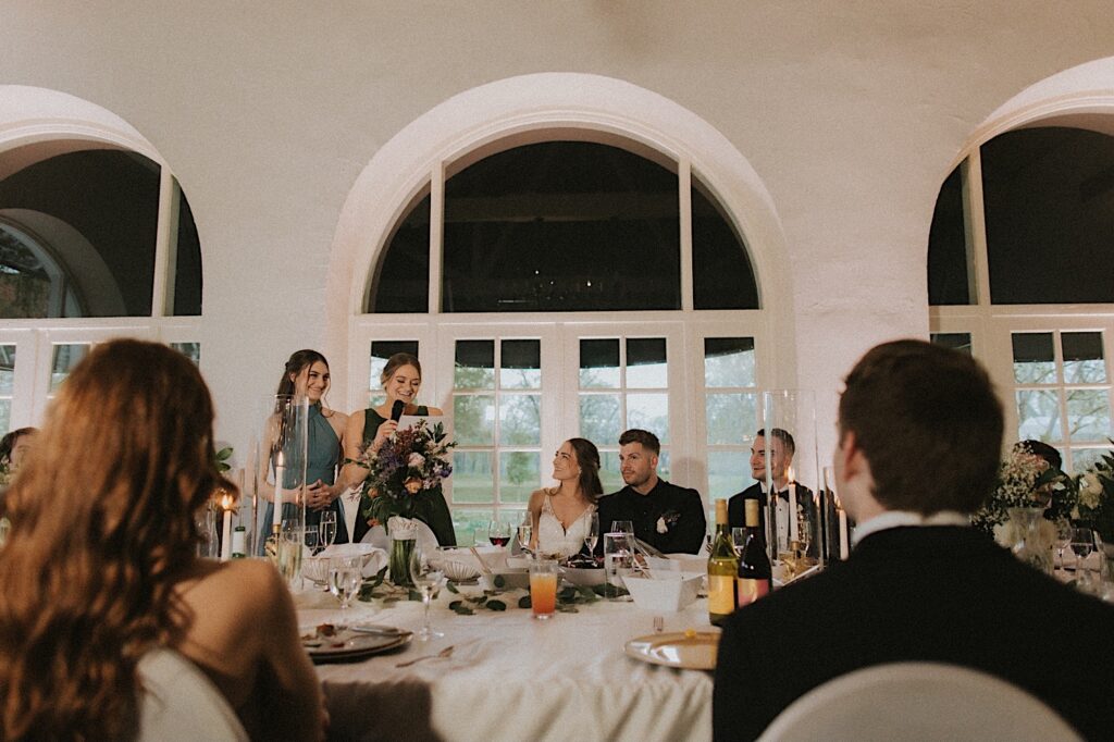 Bride and Groom sit at the head table and smile at guests giving a wedding toast standing next to them. 