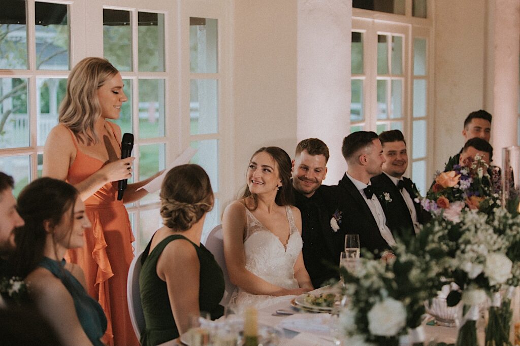 Bride smiles as bridesmaid gives a toast. They are sitting at the head table seated around their bridesmaids and groomsmen. 