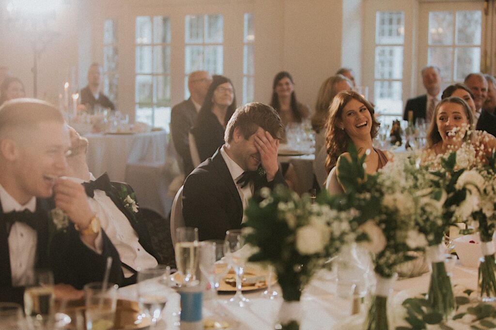 Guests puts his hand on his face laughing as other guests laugh around him. They are seated at gorgeous head table adorned in beautiful white flower bouquets in the reception hall at Ravisloe Country Club. 