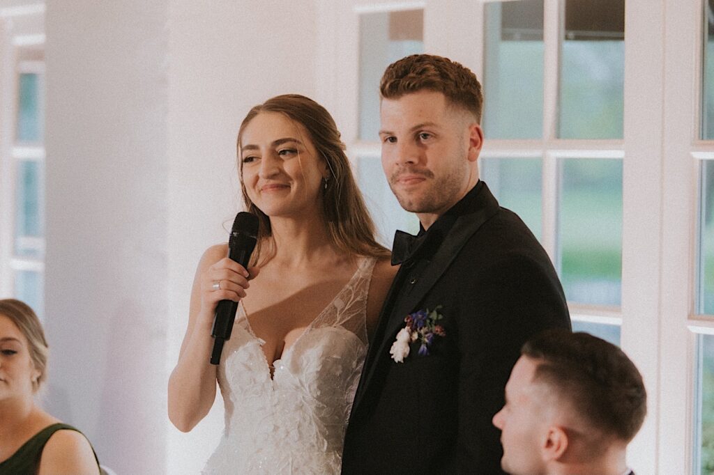 Bride smiles as she holds a microphone standing next to the groom during their reception at Ravisloe Country Club. 