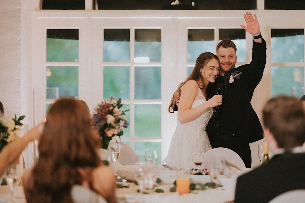 Bride hugs the groom as he puts his hand up and guests seated around them clap. 