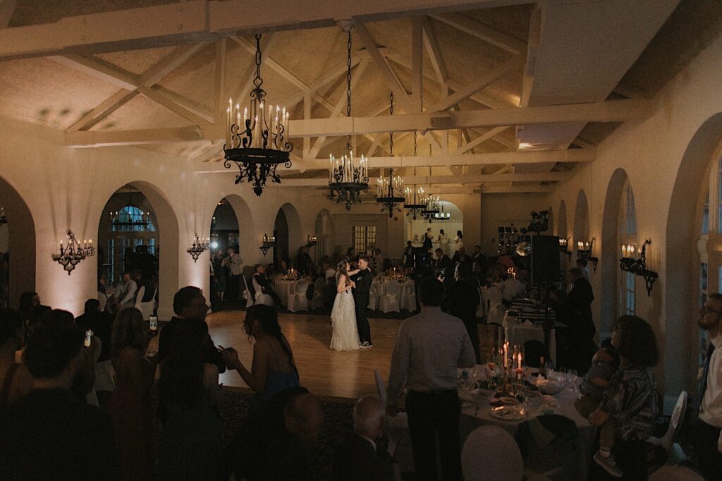 Chicagoland Wedding photographer captures a wide shot of the reception hall at Ravisloe Country Club focusing in on bride and groom during their first dance. From the ceiling hangs beautiful chandeliers and the dance floor is lit on the couple. 