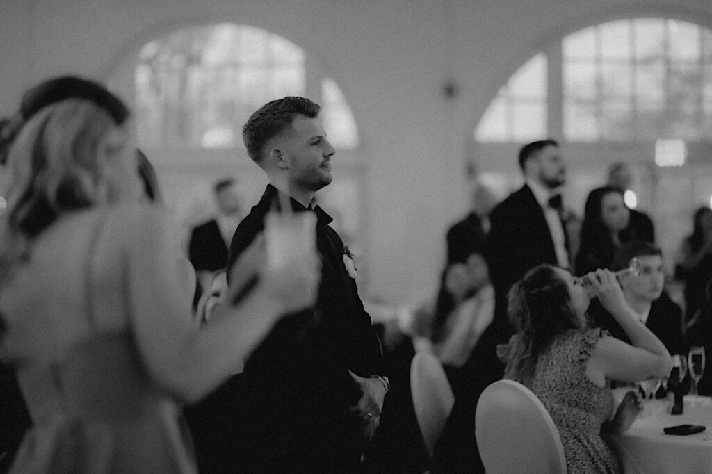 Groom watches the dance floor  surrounded by guests during beautiful reception.  