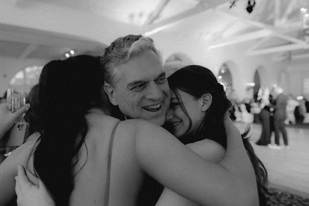 Father of the bride is hugged by bridesmaids during wedding reception. 