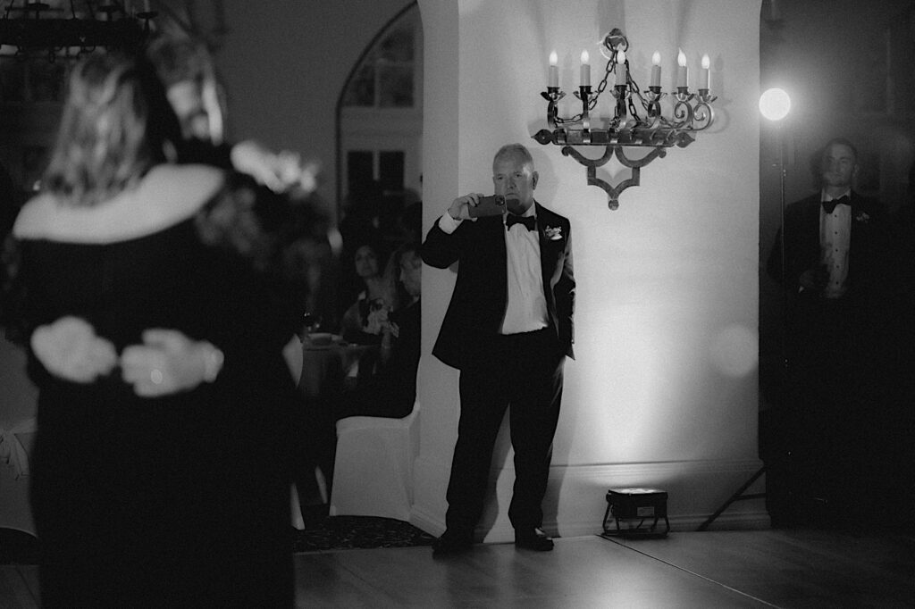 Guest stands next to a chandelier as he videos the groom and his mom during their dance at wedding reception. 