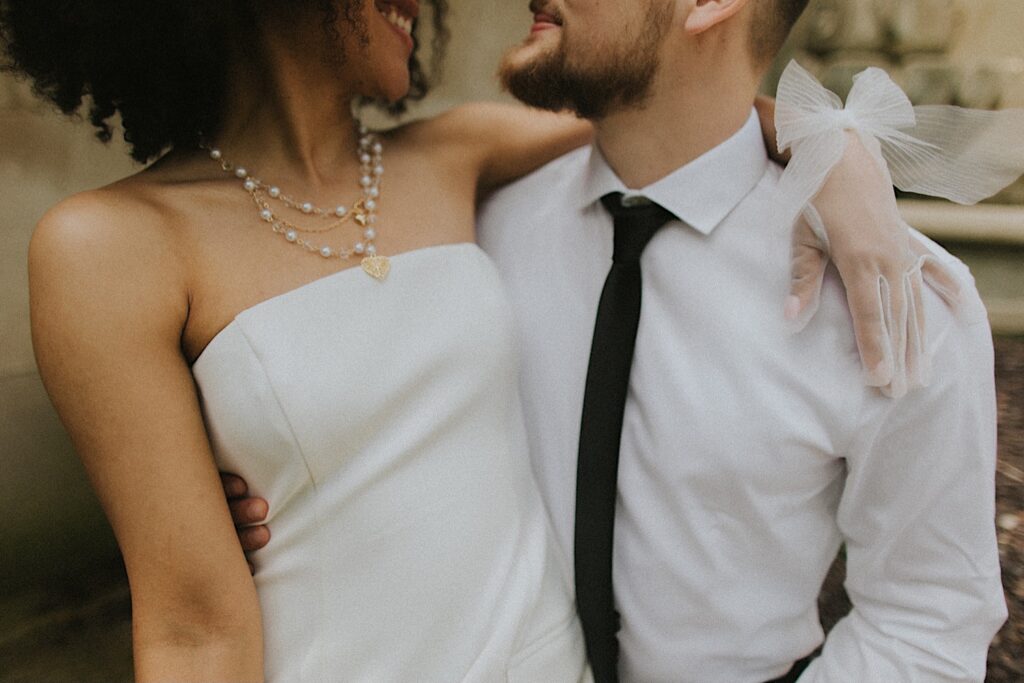 Springfield Elopement Photographer captures bride sitting on Groom's lap. The portrait is captures from their mouths down. The bride is wearing a strapless dress with a layered pearl necklace and heart charm. The groom is wearing a white button up with a black tie.