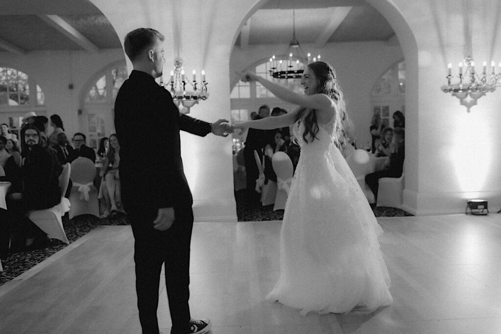 Black and White photo of bride pointing to the crowd as she dances with groom on dance floor surrounded by chandeliers during reception at Ravisloe Country Club. 