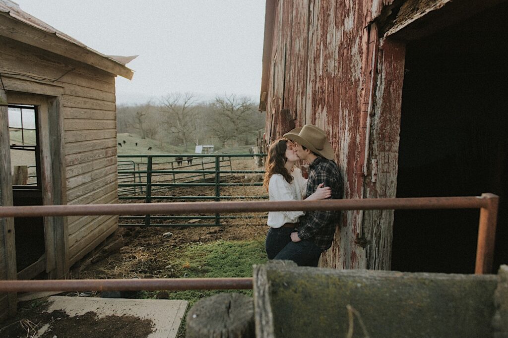 Couple kisses while leaning up against red barn on a farm in Springfield, Illinois for engagement photos. 