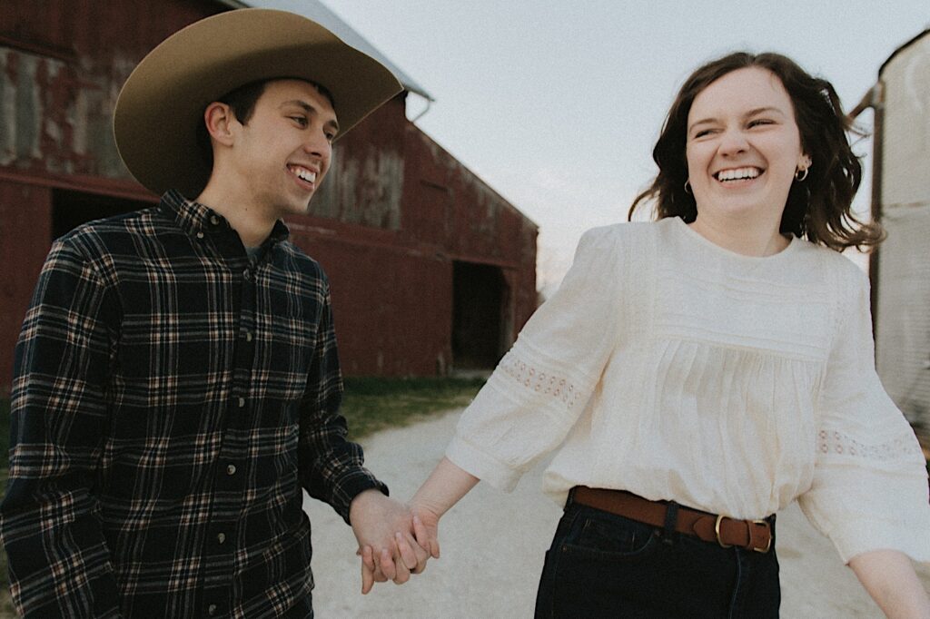 Couple laughs while running between barns during engagement photo session on a farm in Springfield Illinois 