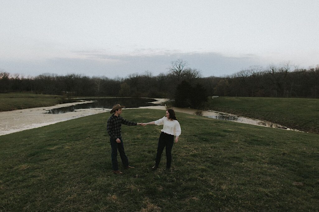 Couple dances surrounded by grass and a pond at dusk for romantic engagement photos taken by Central Illinois Engagement photographer. 