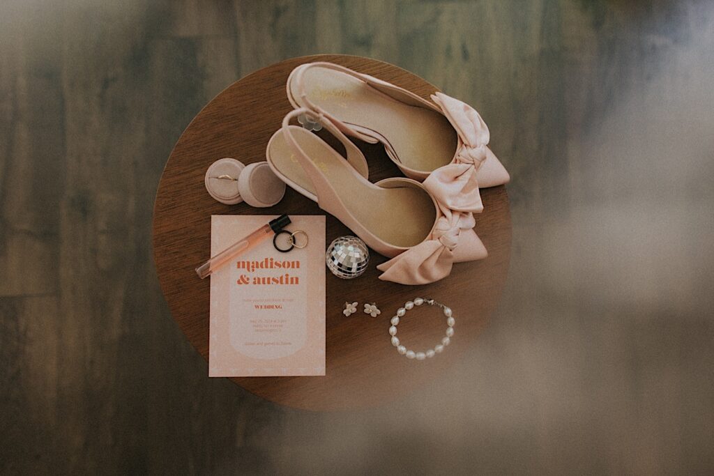 Details of bride's accessories including blush shoes, mini disco ball, ring, pearl bracelet and wedding invitation sitting on a wooden table.