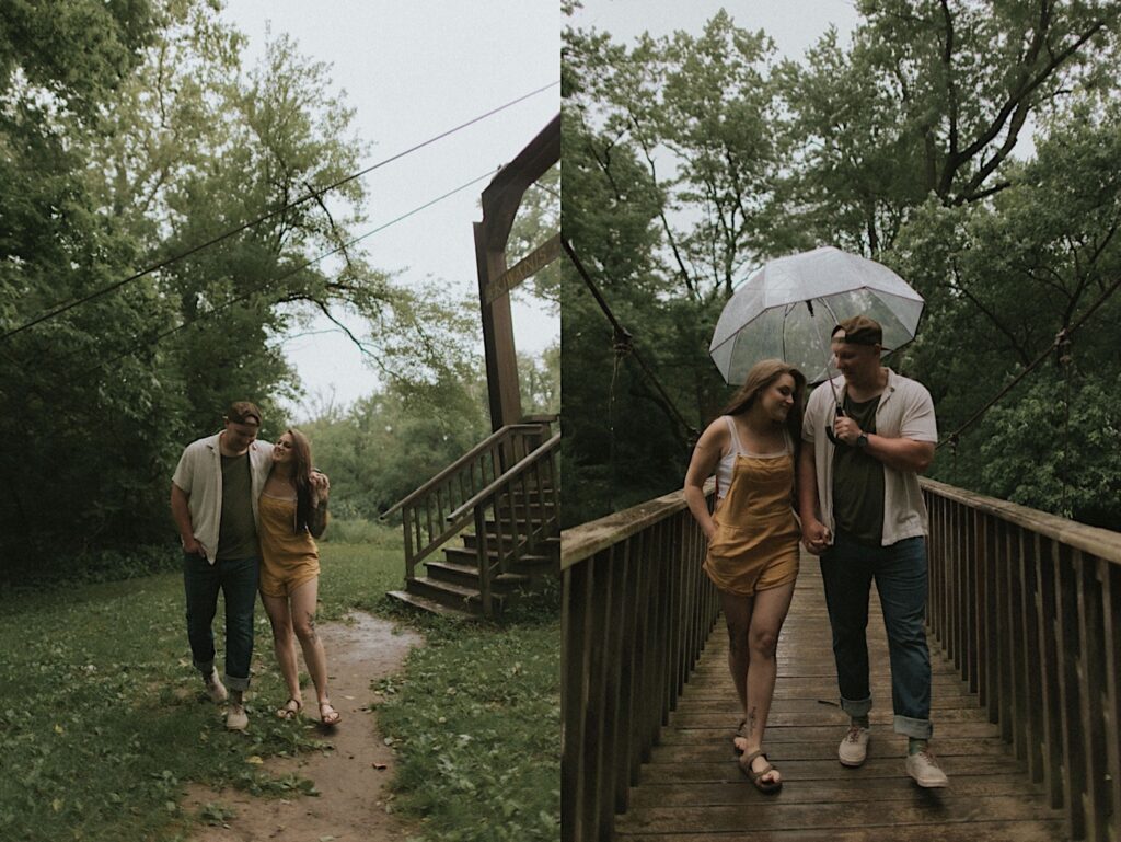 Woman in yellow overalls holds onto her fiancés hand as they walk over a forested bridge holding a clear umbrella. They're walking in the rain for moody engagement photos in Lincoln, Illinois. 