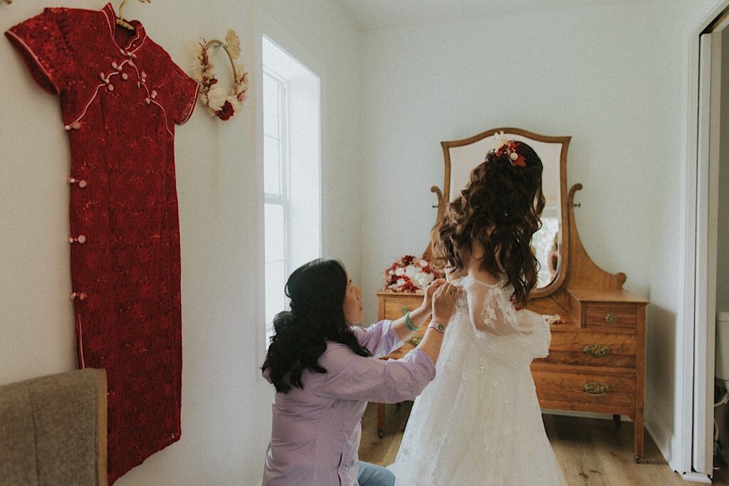 Bride's mother helps button her dress while they stand in front of a beautiful wooden vanity while getting ready for wedding ceremony. 