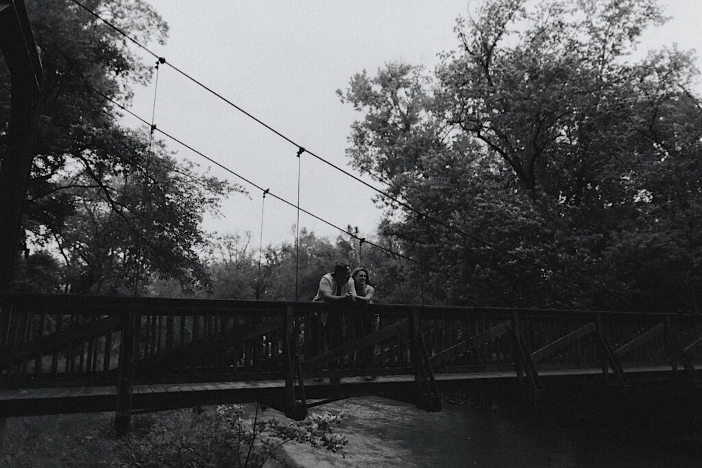 Couple stands on a wooden bridge and puts their elbows on the edge and look out into a stream for a moody black and white engagement portrait. 